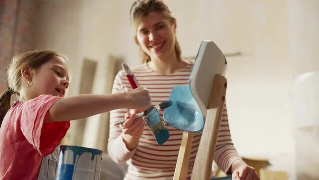 Moving in and Home Renovations: Happy Smiling Mother and Daughter Painting Furniture Chair. Mom Teaches Child to be Artist, Making Apartment Cozy with Art, Color.  Low Angle Portrait Slow Motion