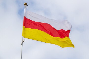 The flag of North Ossetia flies against a clear blue sky with white clouds. Close-up, perfect for news.