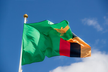 The flag of Zambia flies against a clear blue sky with white clouds. Close-up, perfect for news.