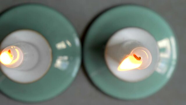Closeup dolly shot of two burning wax candles in green holders, standing next to each other, with focus on the wicks and flickering flames.