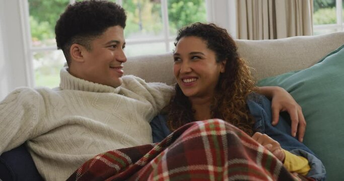 Video of happy biracial couple siting on sofa under blanket