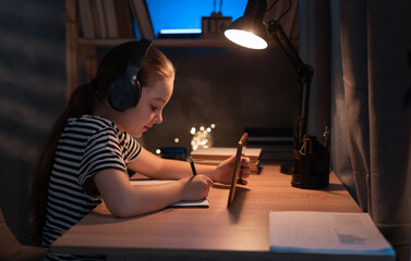 Happy teen girl at digital tablet doing homework online sitting at table at home in dark. Distant education, remote learning