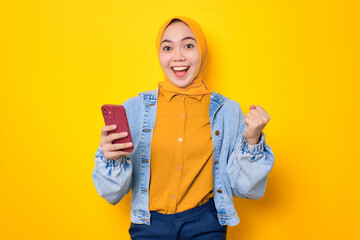 Excited young Asian woman in jeans jacket using mobile phone, celebrating success, getting good news isolated over yellow background
