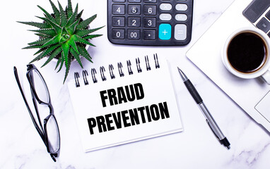 A laptop, glasses, a pen, a calculator, a cup of coffee, a green plant and a white notepad with the text FRAUD PREVENTION lie on a table with task lighting. View from above. Business concept.