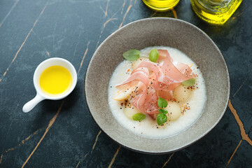 Bowl of cold melon soup served with jamon and fresh basil, high angle view on a dark-olive marble background, horizontal shot