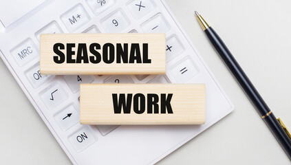 Wooden blocks with the SEASONAL WORK Iie on a light background on a white calculator. Nearby is a black handle. Business concept