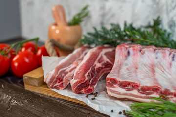 Raw pork loin, bacon with ribs on a wooden board, spice mortar, and pestle, rosemary, tomatoes. Pieces of meat, ingredients for cooking. Protein nutrition, a diet with fresh herbs and vegetables