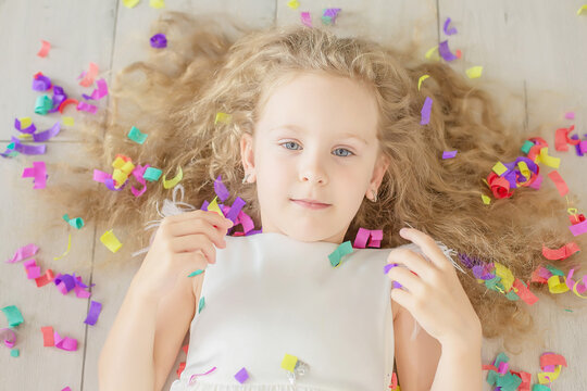 happy cute blonde girl with curly hair toothy smile lies on the floor confetti falls on her face birthday party
