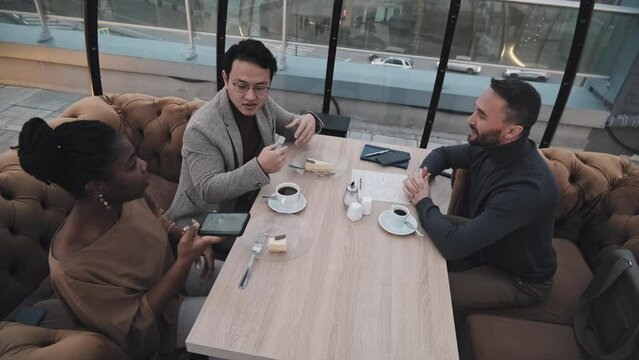High angle of three diverse young friends sitting at table in modern restaurant, talking and taking pictures with smartphone