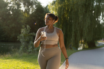 Plus sized African American woman using mobile phone and earphones during the walking at the park in a summer day - obrazy, fototapety, plakaty