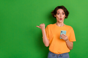 Photo of shiny shocked lady wear orange outfit pointing thumb empty space reading modern gadget isolated green color background