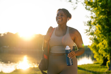 Young African American woman walking with bag and bottle of water through the park in a summer day