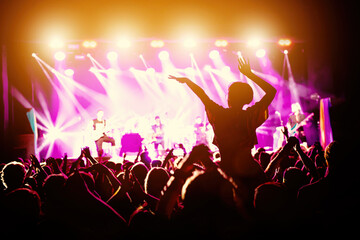 The girl with raised hands during the music concert. Crowd and stage light in a concert hall.