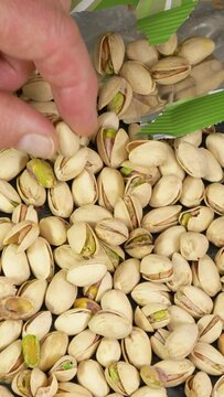 Vertical video social media format – Closeup of a hand taking a roasted, salted pistachio nut / kernel and shell, from the spread out contents of an opened retail packet.