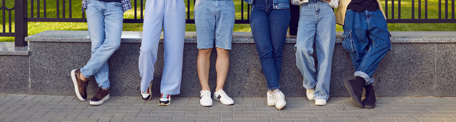 Legs of friends. Legs of six young people in stylish denim clothes standing in row on city street. Horizontal image of male and female legs in different denim pants and shorts. Panoramic banner.