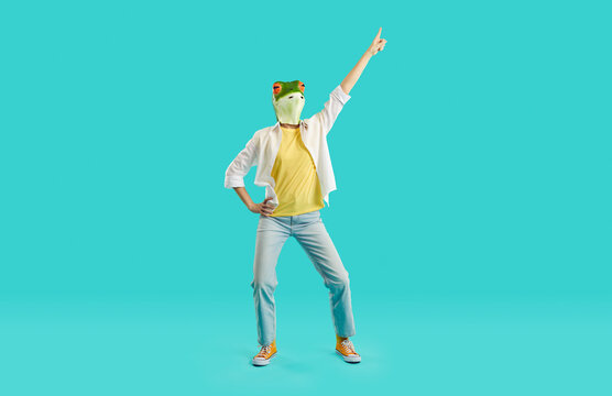 Cheerful Woman With Frog Head Shows Funny Dance Moves Having Fun On Light Blue Background. Unknown Woman In Casual Clothes And With Mask In Shape Of Frog's Head Dances With Her Hand Raised.Full Length