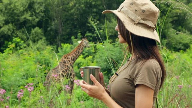 Pretty woman traveler with backpack drinking water while standing on mountain. A girl drink water in sand desert safari. a woman zoologist drinks water from a tourist iron mug and watches a giraffe