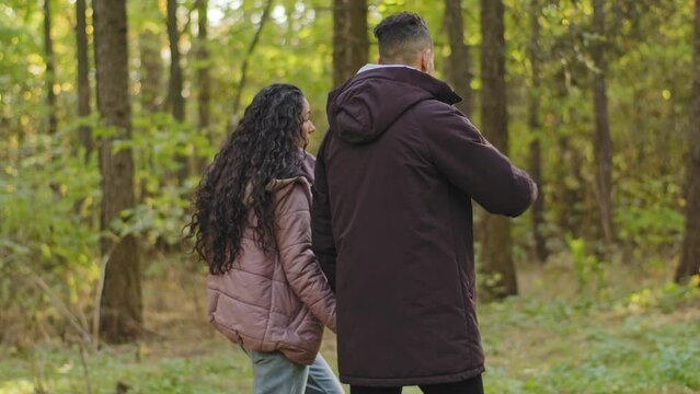 Back view young loving carefree joyful hispanic couple walks in autumn park in nature enjoying weekend together walking outdoors holding hands friendly communicate serious guy explaining to girl talks