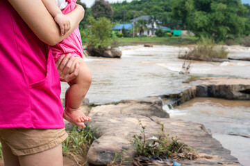A mother is holding an infant baby with outdoor waterfall or cataract as blurred background. People...