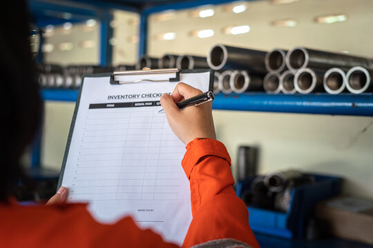 A worker is using pen to writing on inventory checklist form, with blurred background of storage shelf in the factory. Industrial working action photo. Selective focus.	
