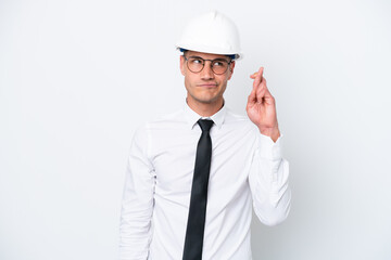 Young architect caucasian man with helmet and holding blueprints isolated on white background with fingers crossing and wishing the best