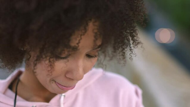 Trendy and stylish young female texting or replying on her phone outdoors. An attractive woman with an afro listening to music using earphones and sending emails in the city on a windy day
