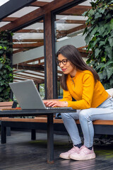 Young hispanic female student with indigenous features wearing a yellow sweater sitting on a bench outside using her laptop