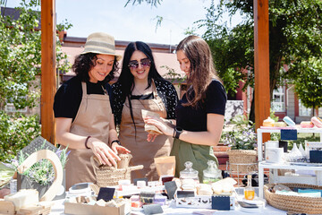 Three brunette women sellers of European appearance in aprons at a street fair laughing discuss their goods - obrazy, fototapety, plakaty