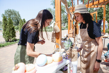 Young brunette girl buyer with long hair and woman seller in apron and hat on either side of the counter with goods at the street fair in summer