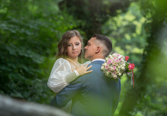 a guy in a suit and a girl in a white dress are standing in the forest and hugging.