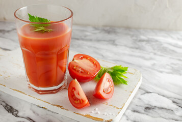 One clear glass cup with red tomato juice and celery leaf, sliced tomato on a white cutting board. Healthy food concept. Horizontal orientation. Selective focus. Cpy space.
