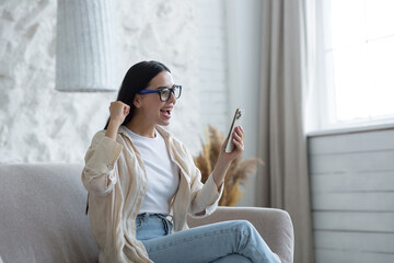 Beautiful young brunette woman wearing glasses at home in living room shocked received good news,...