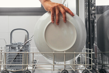 Woman's hands taking out or putting a white plate in the metal tray of the automatic dishwasher. Young girl placing the tray with cutlery and dishes to perform a wash with one of the appliances