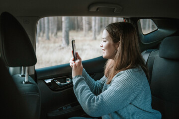 Fototapeta na wymiar Smiling caucasian teen girl in profile with loose hair wearing grey knitted sweater sitting inside the car and taking photo with smartphone through the window