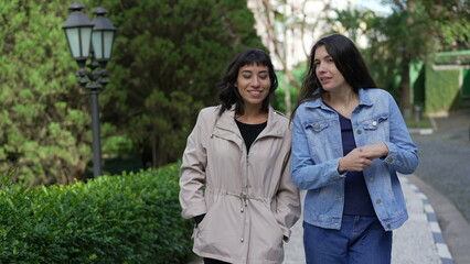 Fototapeta premium Two female friends gossiping while walking outside in street. Young women conversation on sidewalk