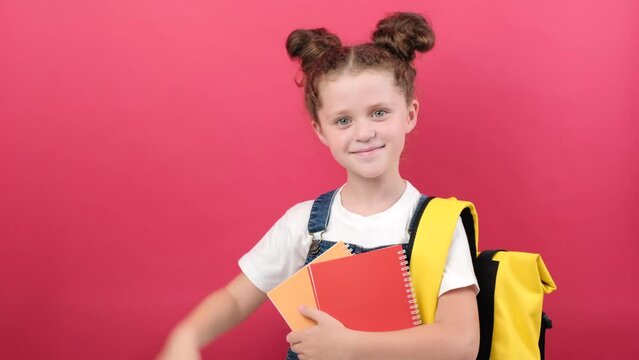 Portrait of school kid girl 6-7 years old in white t-shirt and yellow backpack holding two notebooks pointing fingers on workspace isolated over red background studio. Education lifestyle concept