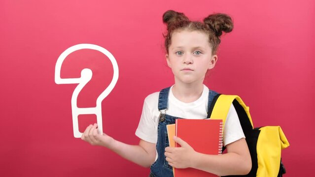 Pensive little kid girl holding big question marks and notebooks, wears t-shirt and yellow backpack, posing isolated over pastel red color background wall in studio. Education and school concept