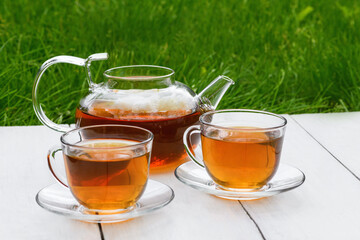 Tea in glass teapot and two cups on a wooden white background background against the background of green grass. Teapot with green or black tea. Tea time