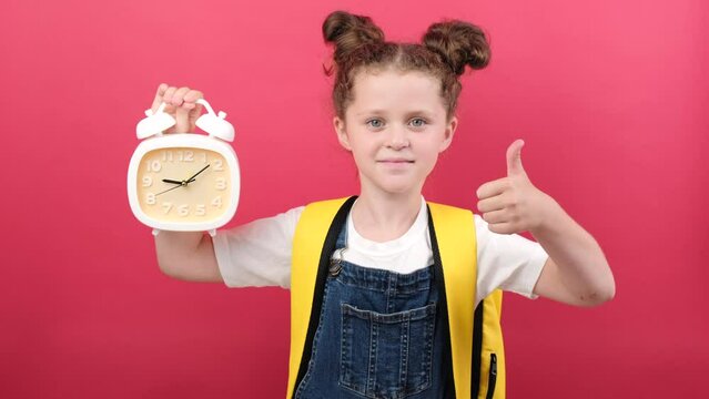 Smiling little kid girl 6-7 years old in white t-shirt hold clock showing thumb up, posing isolated on pastel red color background wall in studio. Childhood lifestyle concept. Mock up copy space