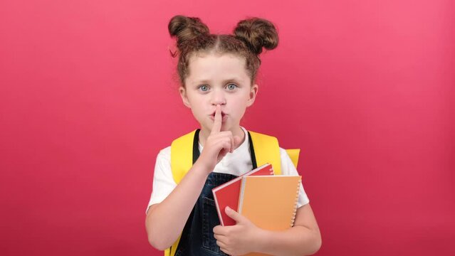 Secret little smart kid school girl with yellow backpack hold books secret say hush be quiet, finger on lips shhh gesture, posing isolated on pastel red studio background. Childhood education concept
