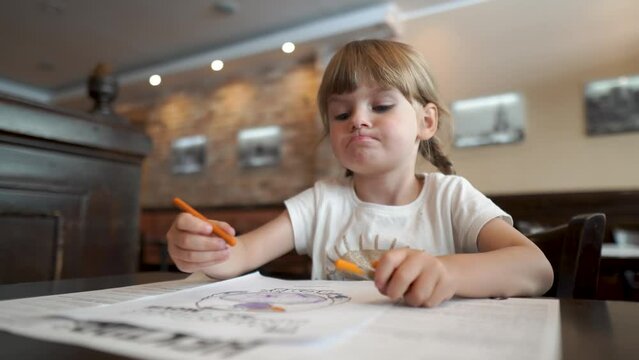A beautiful little girl emotionally coloring a picture in a cafe. Everyday development of children. Leisure of the daughter while waiting. Facial expressions and grimaces.