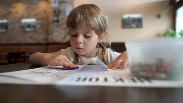 Little beautiful girl coloring a picture in a cafe. Everyday development of children. Leisure of the daughter while waiting.