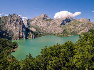 Paisaje de montaña con un lago alpino rodeado de bosques y montañas rocosas con un cielo azul de nubes blancas.