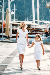 Smiling caucasian blonde  mother and daugher walking by harbor with yachts on a touristic sea resort