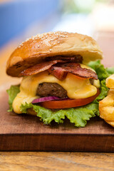 Cheeseburger with cheese, lettuce, tomato, onion, bacon and french fries, served on a wooden board.
