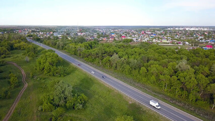 Fototapeta na wymiar Top view of highway passing through village with forest. Clip. Track with passing cars on background of village with green forest and horizon with sky