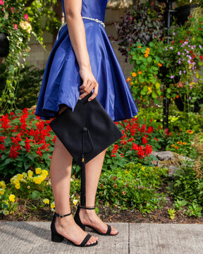 Girl Holding A Graduation Cap Wearing A Royal Blue Dress And Matching Shoes In A Garden.