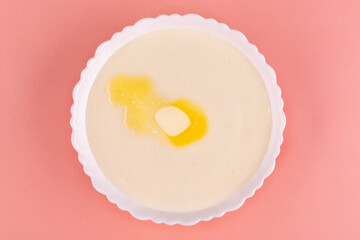 Semolina porridge with a piece of butter in a beautiful white plate (bowl) on a peach-pink background close-up.