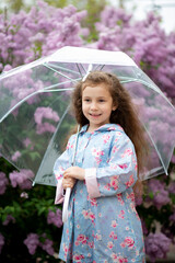 A sweet girl of five years with a transparent umbrella against the background of blooming lilacs enjoys spring and has a fun. Pastel colors. Spring. Childhood. Beauty.