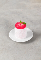 Vegan cherry cream dessert of cylindrical shape with turbillon jelly on top, sprinkled with white coconut chips, on a plate. Light grey background